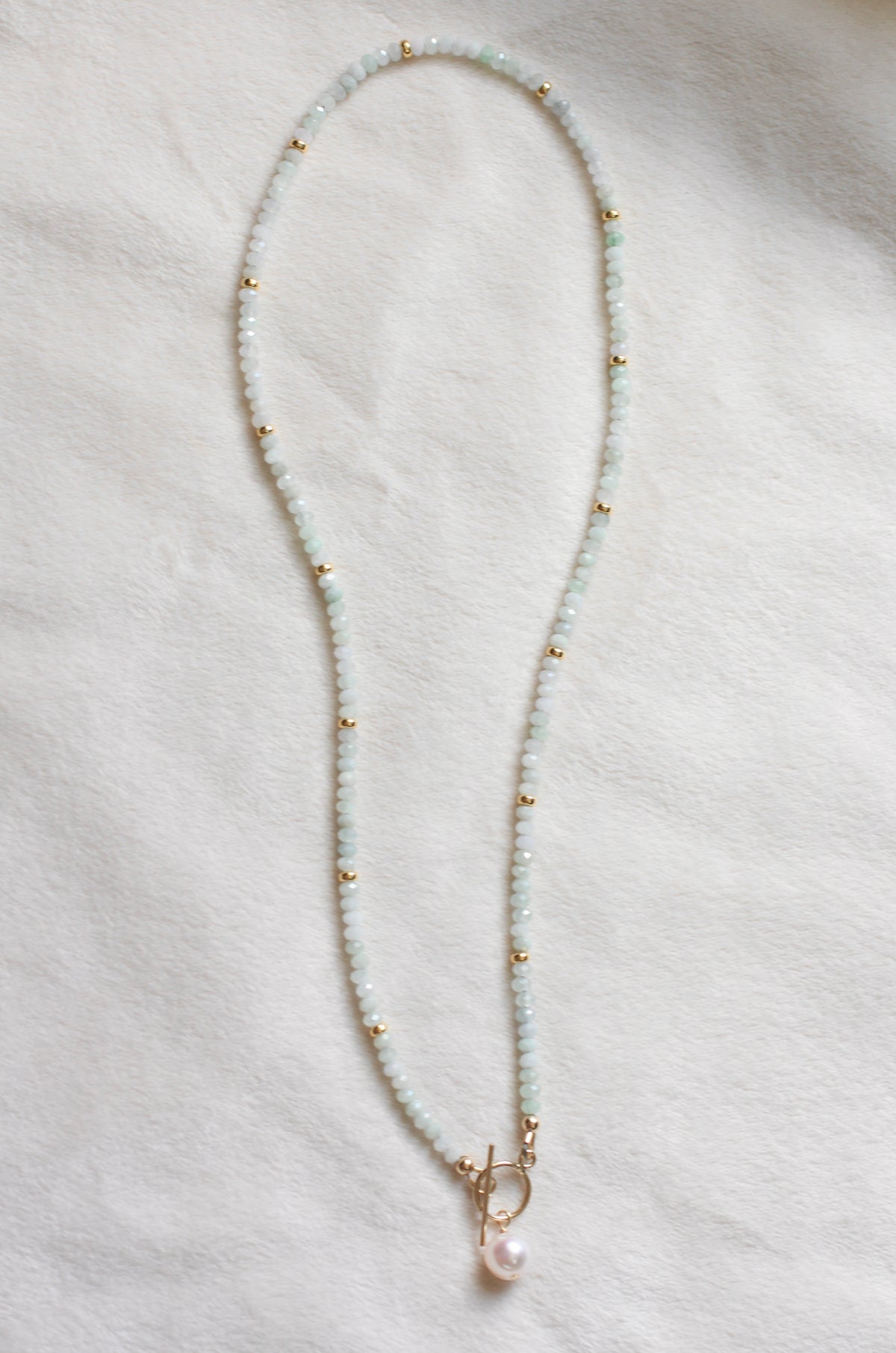 A delicate beaded necklace featuring pale greenish-white round beads, small gold spacer beads, and a gold clasp with a dangling pearl, laid flat on a textured white fabric background.