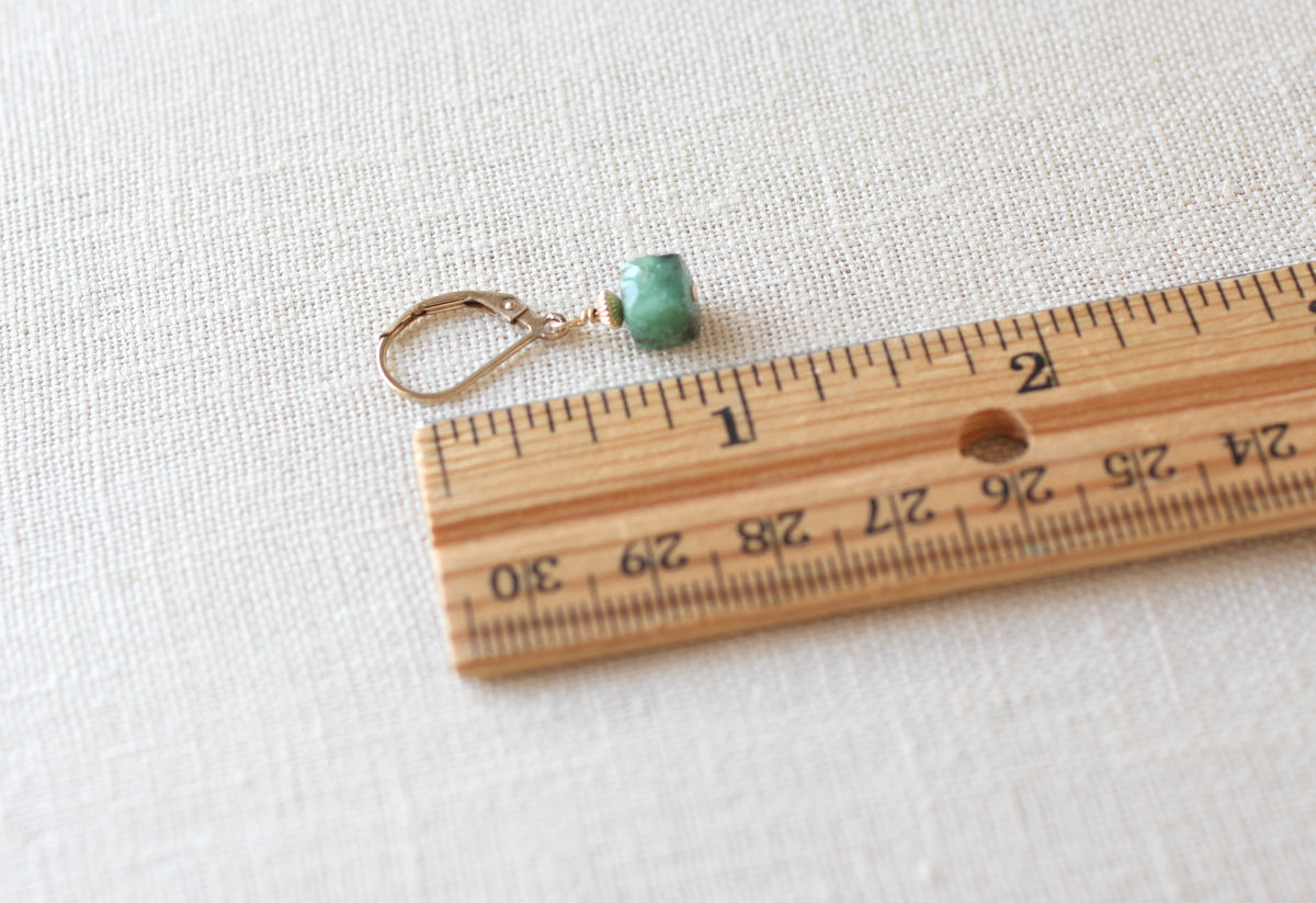 A single drop earring with a small green bead and gold-toned metal, shown next to a wooden ruler on a light fabric background.