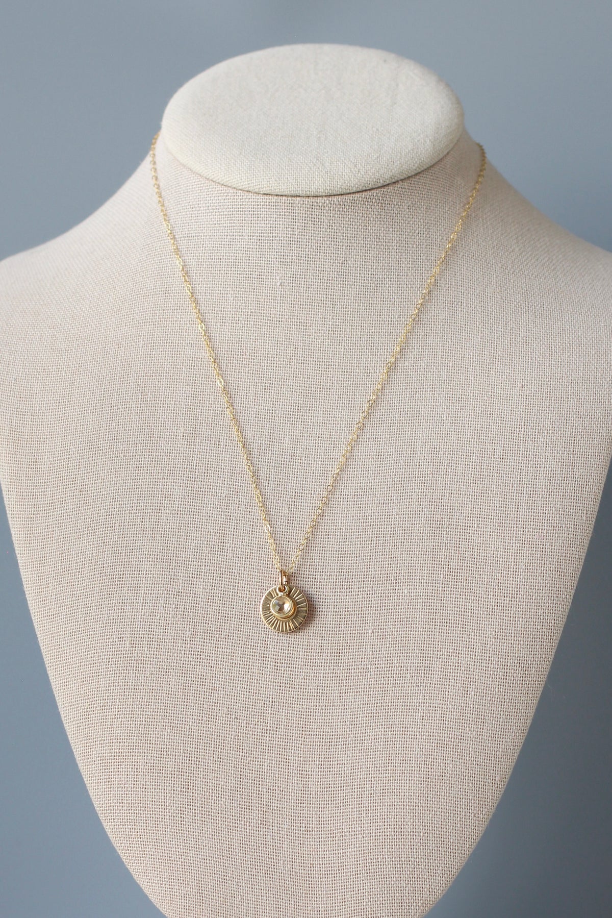 A delicate gold chain necklace with a small sunburst pendant featuring a central clear stone, displayed on a cream-colored fabric jewelry bust against a soft gray background.