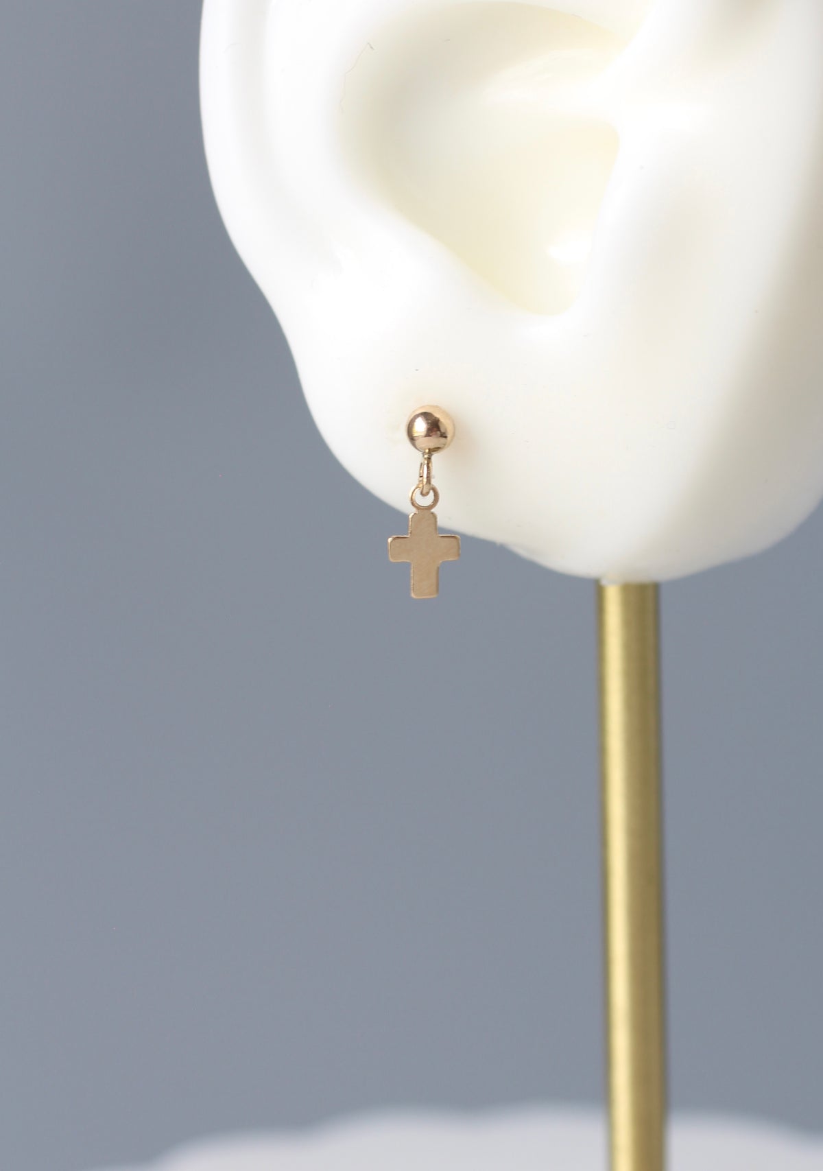A small gold cross dangle stud earring displayed on a white ear mannequin against a soft gray background.