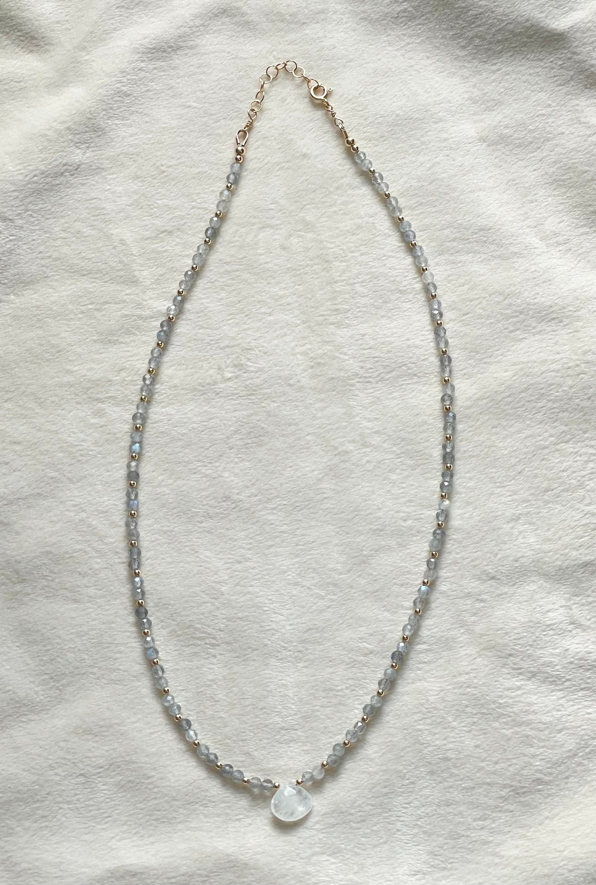 A delicate beaded necklace with small bluish gray faceted beads and a small white teardrop pendant, laid flat on a textured off-white fabric background.