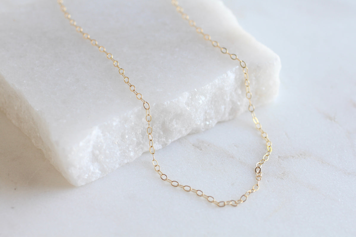 A delicate gold chain necklace draped over a white textured stone slab on a light marble surface.