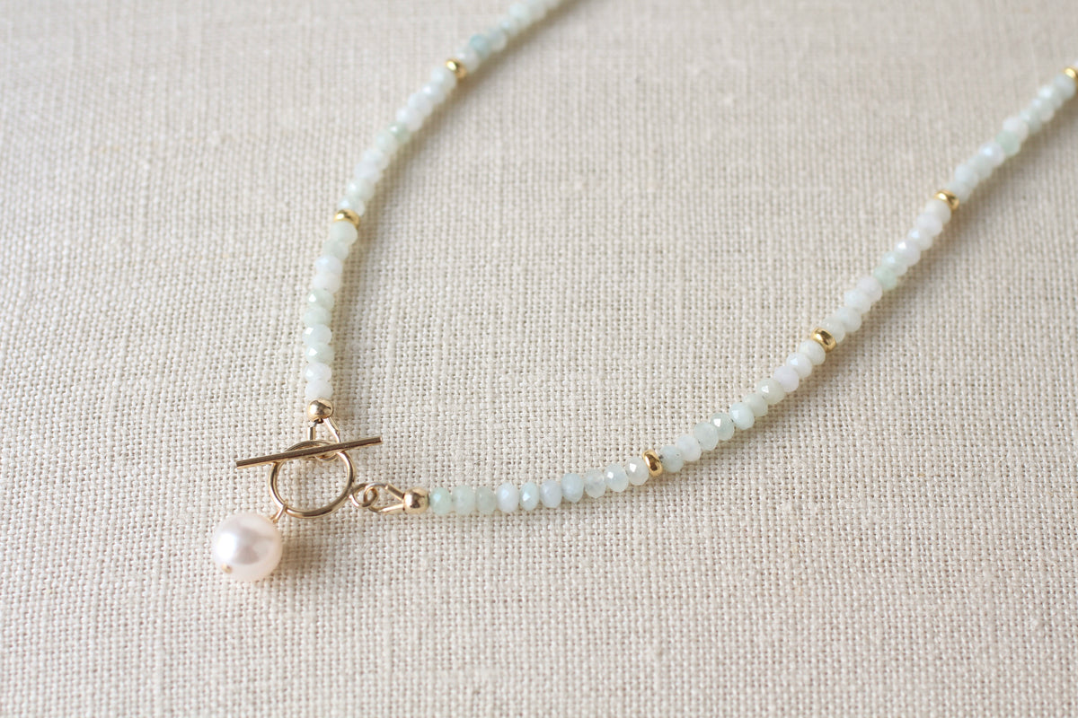 A delicate beaded necklace with pale blue green jade beads, small gold spacer beads, a gold toggle clasp, and a single white pearl pendant, displayed on a textured beige linen background.