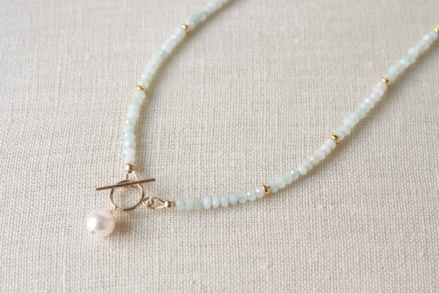 A delicate beaded necklace with pale blue green jade beads, small gold spacer beads, a gold toggle clasp, and a single white pearl pendant, displayed on a textured beige linen background.