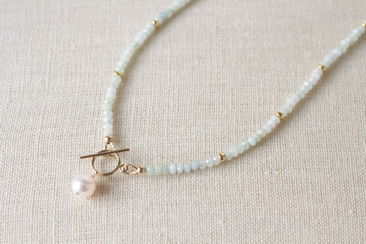 A delicate beaded necklace with pale blue green jade beads, small gold spacer beads, a gold toggle clasp, and a single white pearl pendant, displayed on a textured beige linen background.