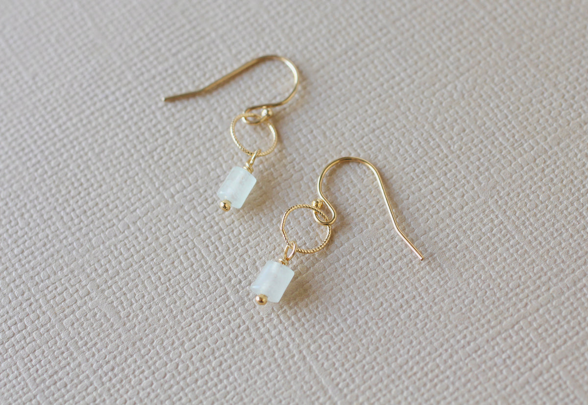 A pair of gold dangle earrings with small rectangular milky light green beads, displayed on a textured beige fabric background.
