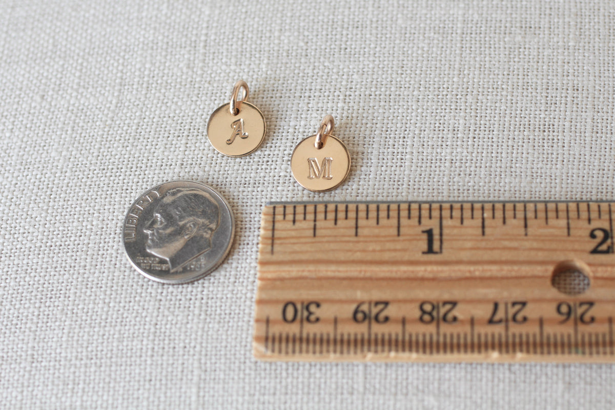 Two small round gold initial charms engraved with letters 'A' and 'M' shown next to a US quarter coin and a wooden ruler on a textured white fabric background.