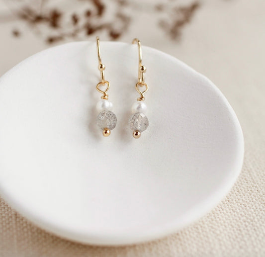 A pair of gold-tone dangle earrings with white pearl beads and translucent gray faceted beads, displayed on a small white dish over a beige fabric background.