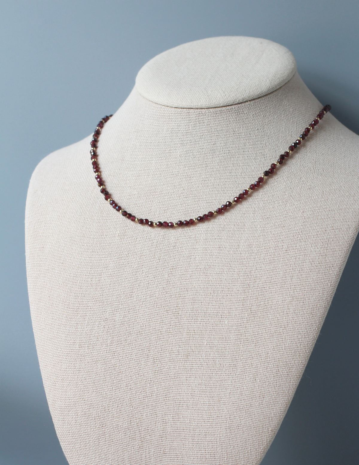 A delicate beaded necklace made of small faceted dark red garnet-like beads, displayed on a cream linen jewelry bust against a soft blue-gray background.