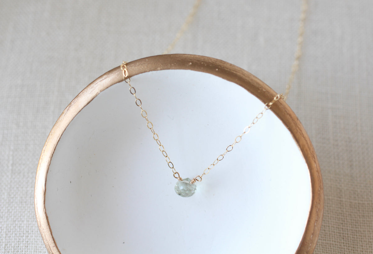 A delicate gold chain necklace with a small round light blue gemstone pendant, draped over a white ceramic dish with a gold rim on a textured beige fabric background.