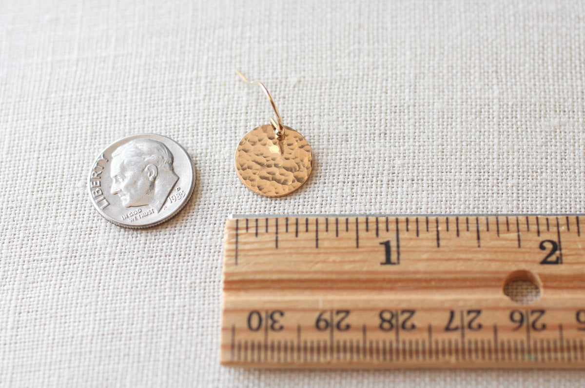 A small hammered round gold earring shown next to a dime and a wooden ruler on a textured off-white fabric background.