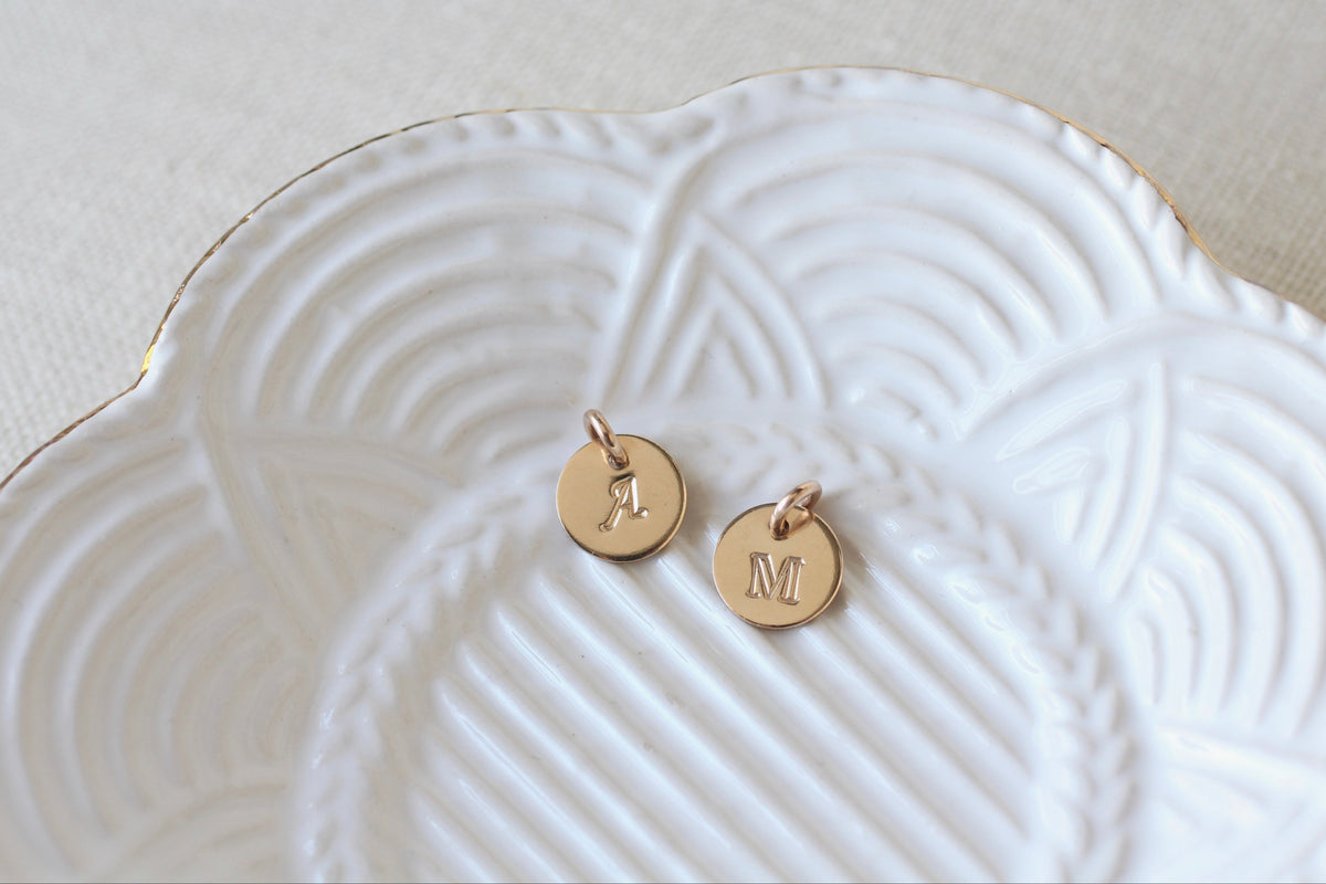 Two small round gold initial charms, one engraved with the letter 'A' and the other with 'M', displayed on a white scalloped ceramic plate with gold trim.
