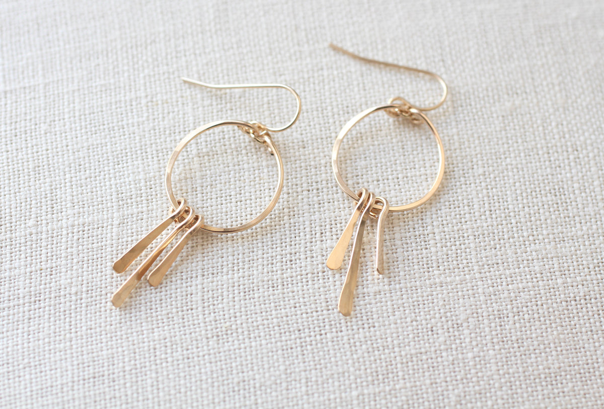A pair of gold-toned dangle earrings with circular hoops and three thin bar charms, displayed on a textured off-white linen background.