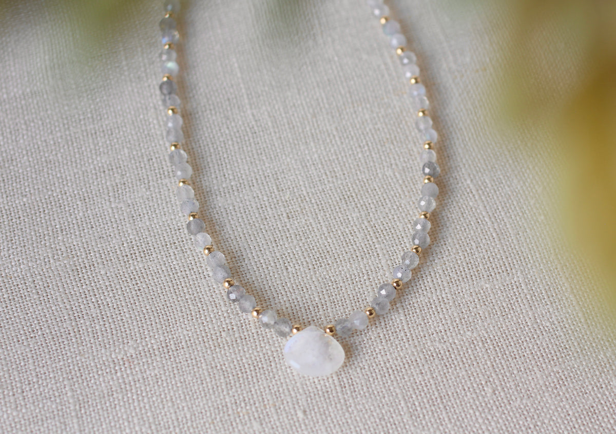 A delicate beaded necklace with small white and gray faceted beads separated by tiny gold spacers, featuring a smooth white teardrop-shaped stone pendant, displayed on a textured beige linen fabric.