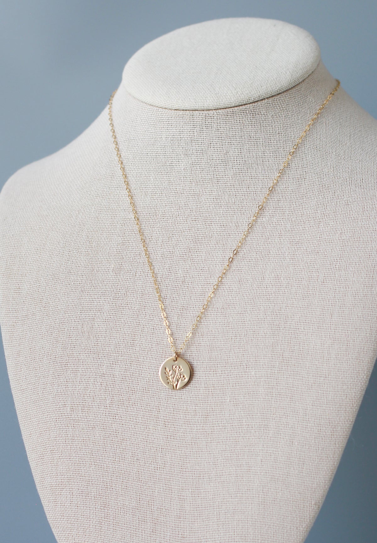 A delicate gold chain necklace with a small round pendant displayed on a cream fabric jewelry bust against a soft blue-gray background.
