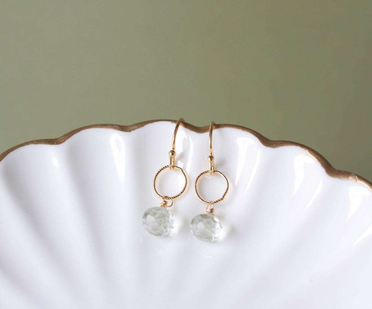 A pair of gold drop earrings with textured circular hoops and faceted clear gemstone pendants, displayed on a white scalloped dish against a muted sage green background.