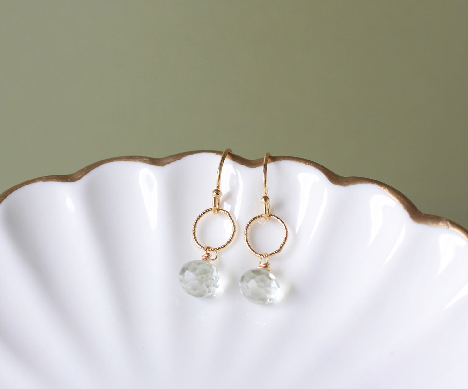 A pair of gold drop earrings with textured circular hoops and faceted clear gemstone pendants, displayed on a white scalloped dish against a muted sage green background.