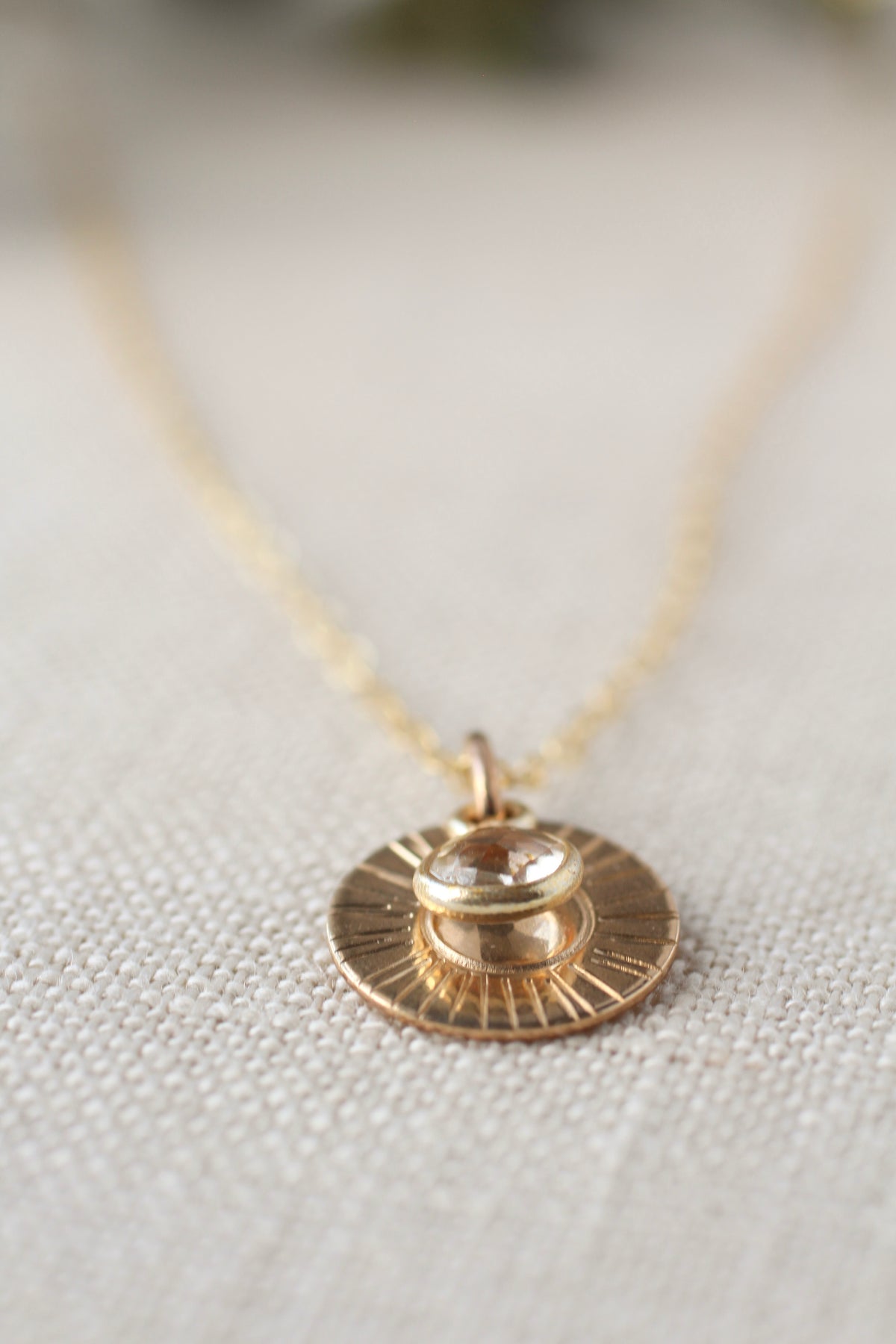A gold necklace with a circular sunburst pendant and a small clear faceted gemstone, resting on a textured beige fabric background.