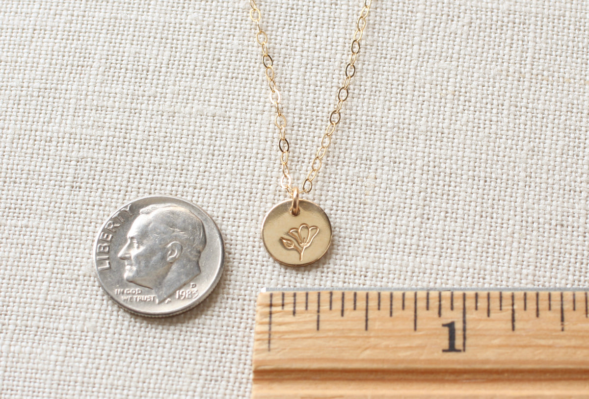 A gold chain necklace with a small round gold disc pendant engraved with a flower, displayed on a textured beige fabric next to a US dime for scale and a wooden ruler.
