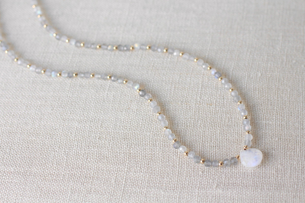 A delicate beaded necklace featuring light gray faceted labradorite beads, small gold spacer beads, and a smooth white oval pendant, displayed on a textured beige linen background.