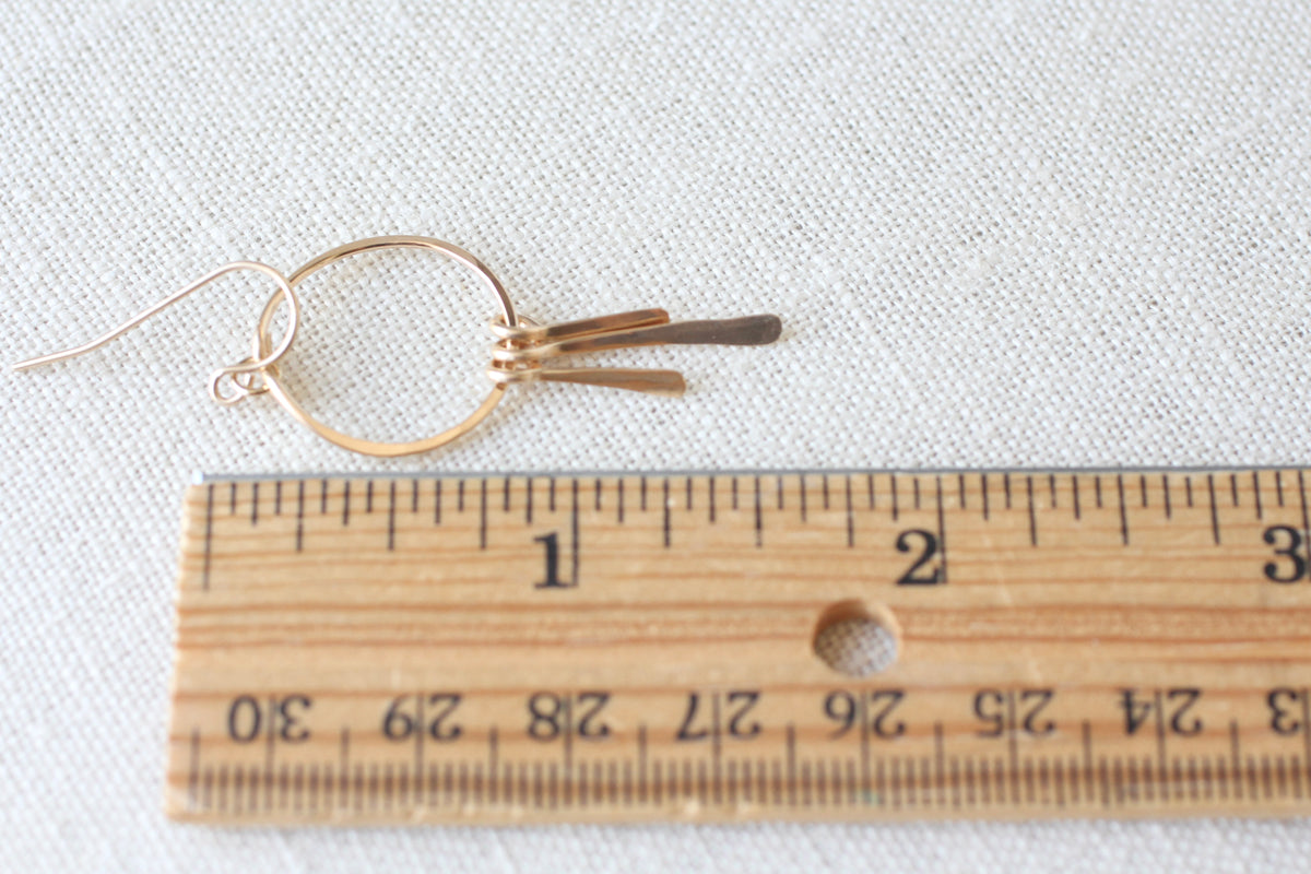 A pair of small gold-toned hoop earrings with three dangling bar elements, shown next to a wooden ruler on a light fabric background.