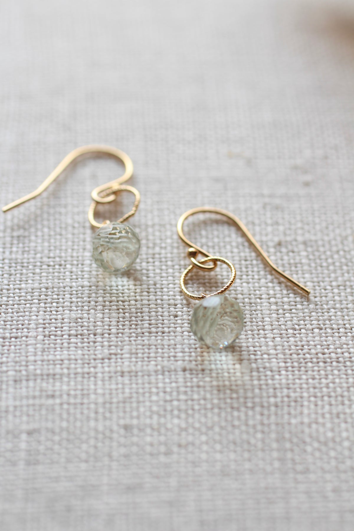 A pair of gold dangle earrings with small, round, textured clear beads, displayed on a neutral beige linen fabric background.