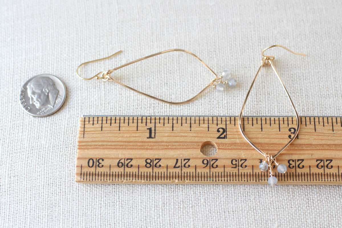 Gold wire hoop earrings with small light blue gray bead drops, shown next to a wooden ruler and a coin for scale on a light fabric background.