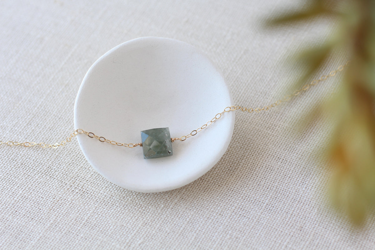 A delicate gold chain necklace with a square-cut green gray gemstone pendant, displayed on a small white ceramic dish over a textured beige fabric background.