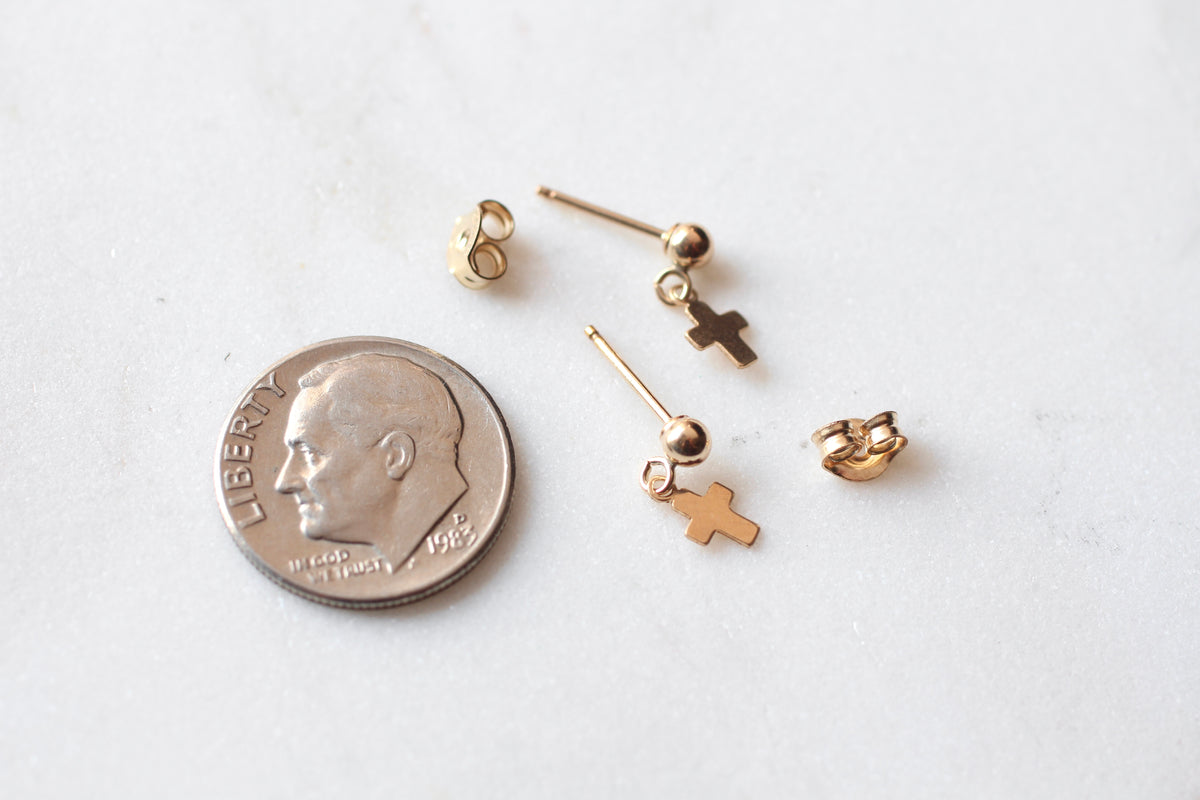 A pair of tiny gold stud earrings with cross charms and ball accents, displayed next to a U.S. dime