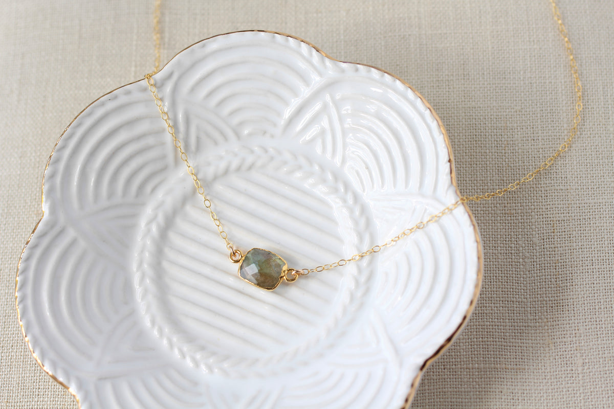 A delicate gold chain necklace with a small, faceted labradorite stone pendant, displayed on a white ceramic dish with gold trim over a beige linen background.