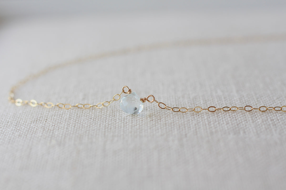 A delicate gold chain necklace featuring a small, faceted light green gemstone pendant, resting on a textured beige linen fabric background.