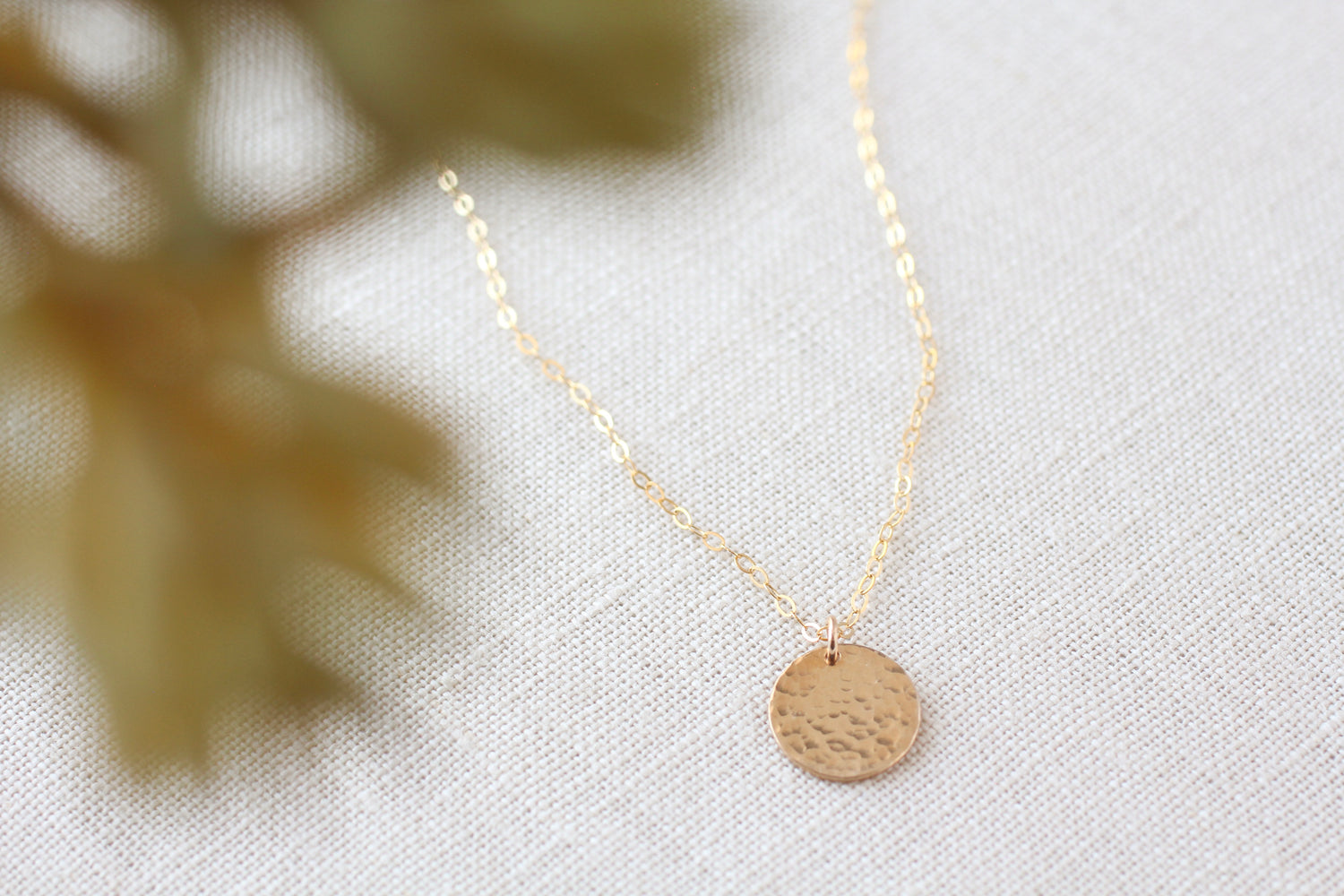A delicate gold chain necklace with a small, textured circular pendant, displayed on a neutral beige linen fabric background with a soft-focus decorative leaf in the foreground.
