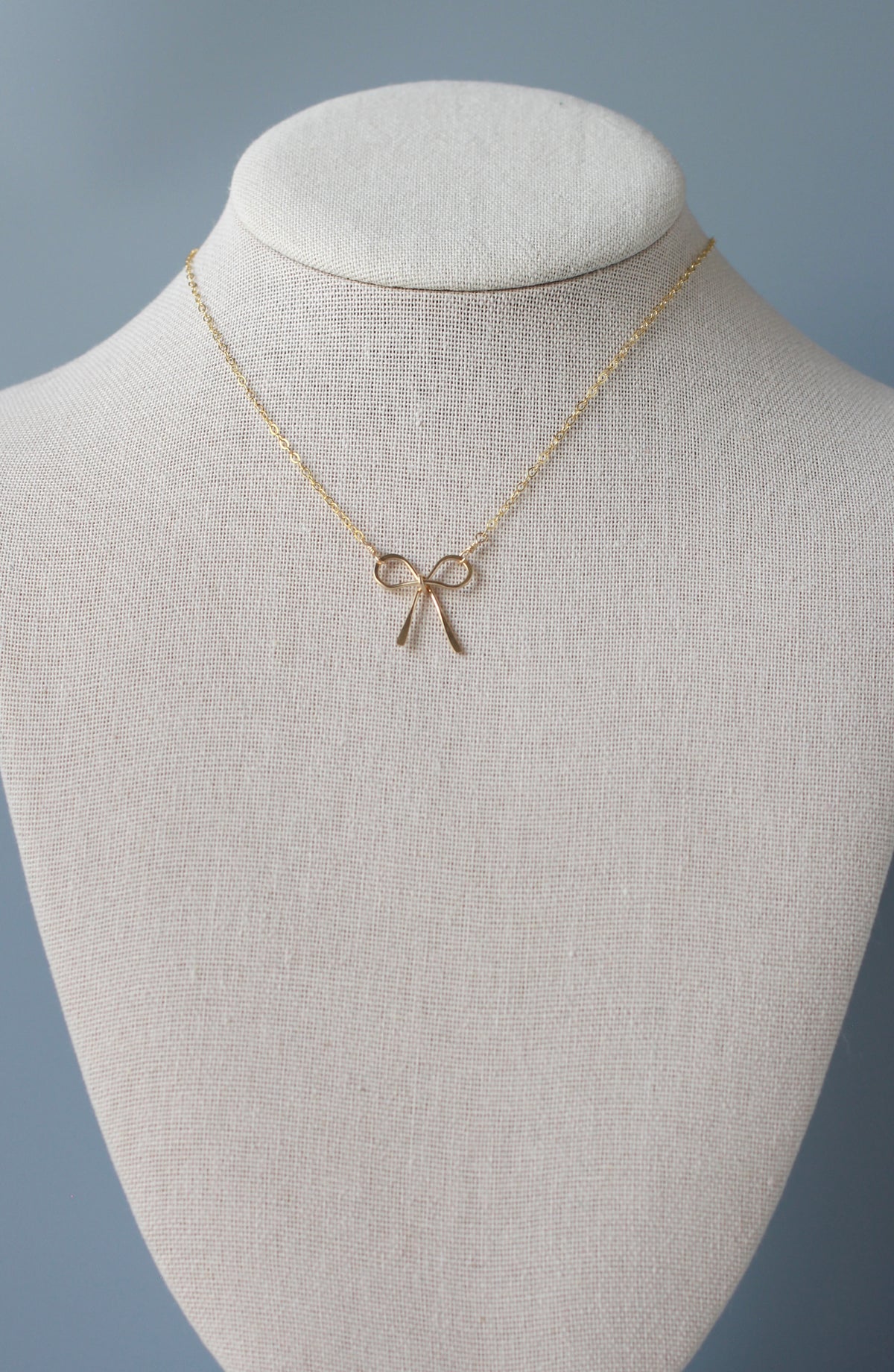 A delicate gold bow pendant necklace displayed on a textured cream fabric jewelry bust against a soft blue-gray background.