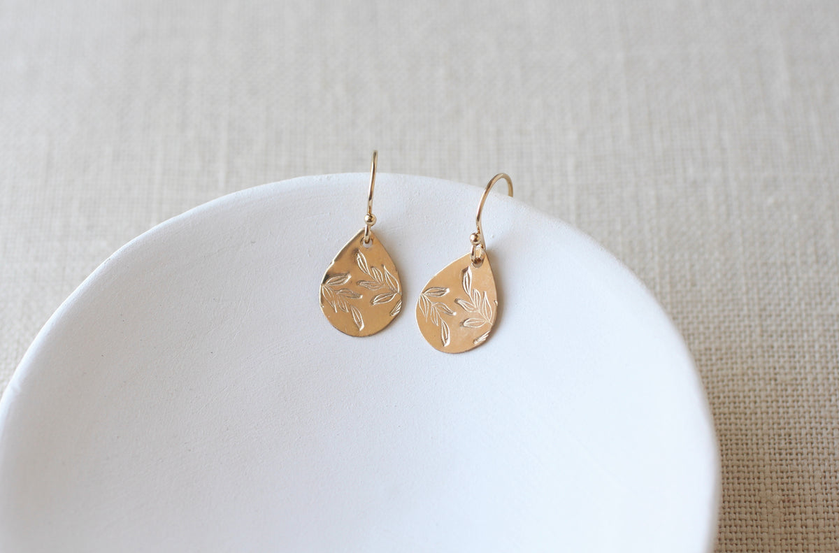 A pair of gold teardrop dangle earrings with engraved leaf patterns, resting on a white ceramic dish over a beige linen background.