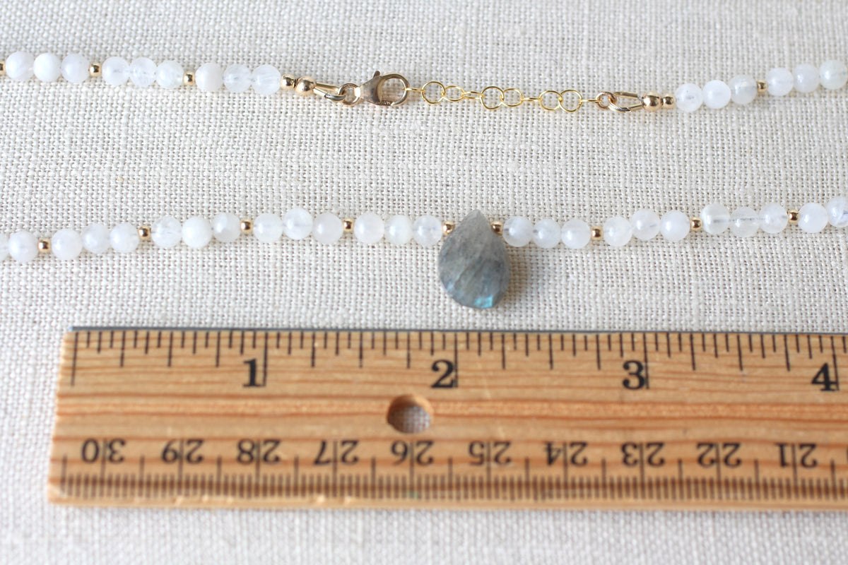 A beaded necklace with white round beads, gold-tone chain links, and a teardrop-shaped gray stone pendant, with a lobster style clasp, shown on a textured beige fabric background with a wooden ruler for scale.