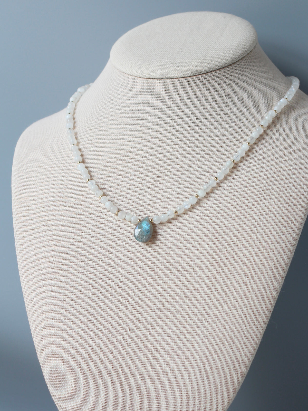 A delicate beaded necklace with small white beads and a central teardrop-shaped blue-green stone pendant, displayed on a beige fabric jewelry bust against a soft gray background.
