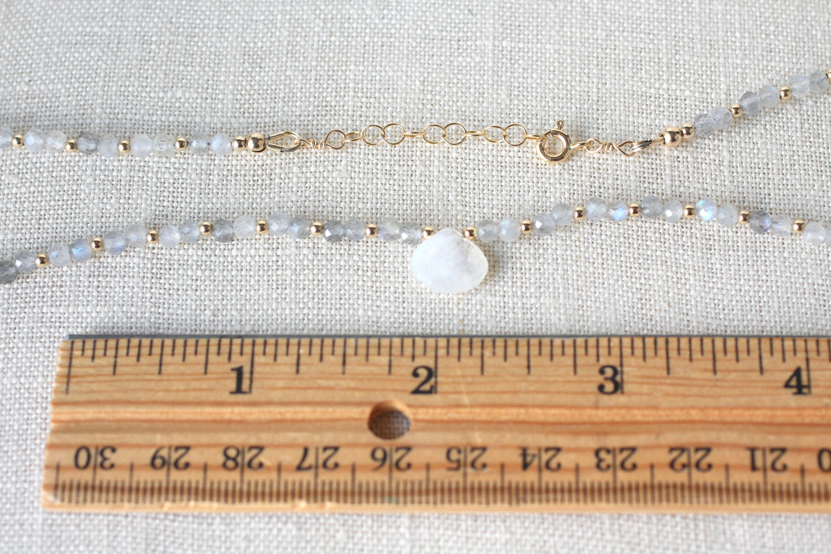 A beaded necklace with pale gray and white round beads, small gold spacer beads, and a white teardrop-shaped stone pendant, shown on a textured beige fabric with a wooden ruler for scale.