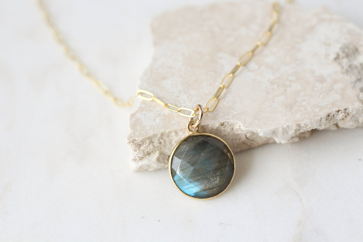 A gold chain necklace with a round bezel-set labradorite pendant, resting on a textured light stone against a white marble background.