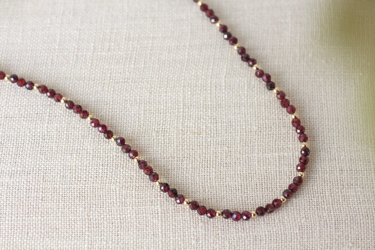 A delicate beaded necklace made of faceted dark red beads separated by small gold spacer beads, displayed on a textured beige linen fabric background.