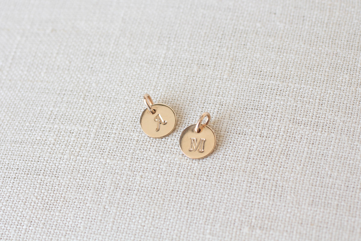 Two small round gold initial pendants engraved with the letters 'J' and 'M', displayed on a textured beige linen fabric background.