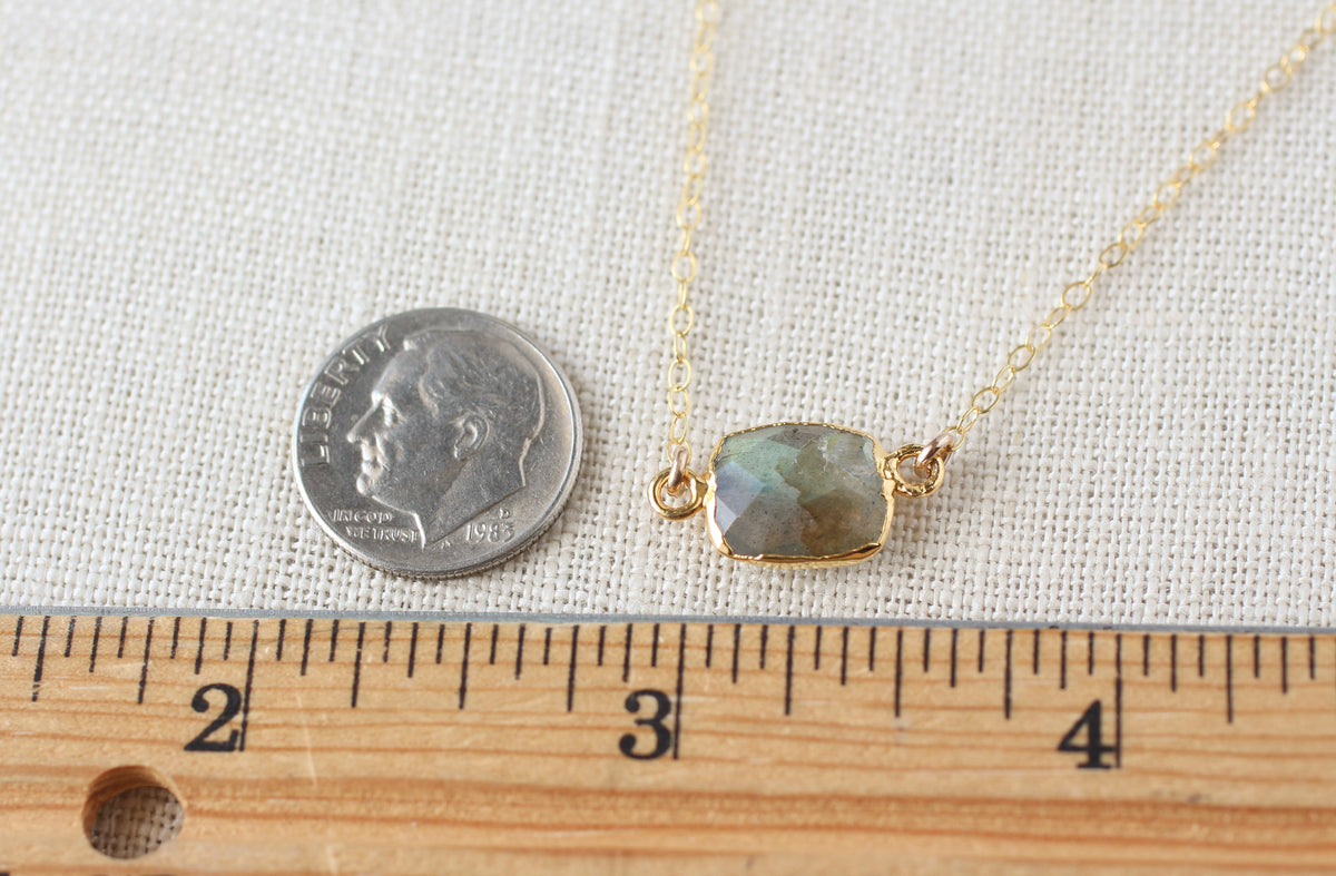 A gold chain necklace with a rectangular iridescent stone pendant, shown next to a U.S. dime