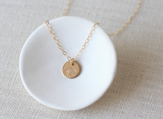 A delicate gold chain necklace with a small round gold disc pendant featuring an engraved floral design, displayed on a white ceramic dish over a beige linen background.