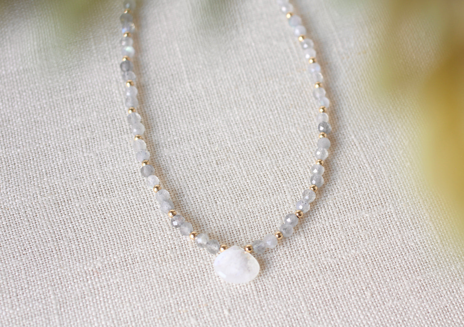 A delicate beaded necklace with small white and gray faceted beads separated by tiny gold spacers, featuring a smooth white teardrop-shaped stone pendant, displayed on a textured beige linen fabric.