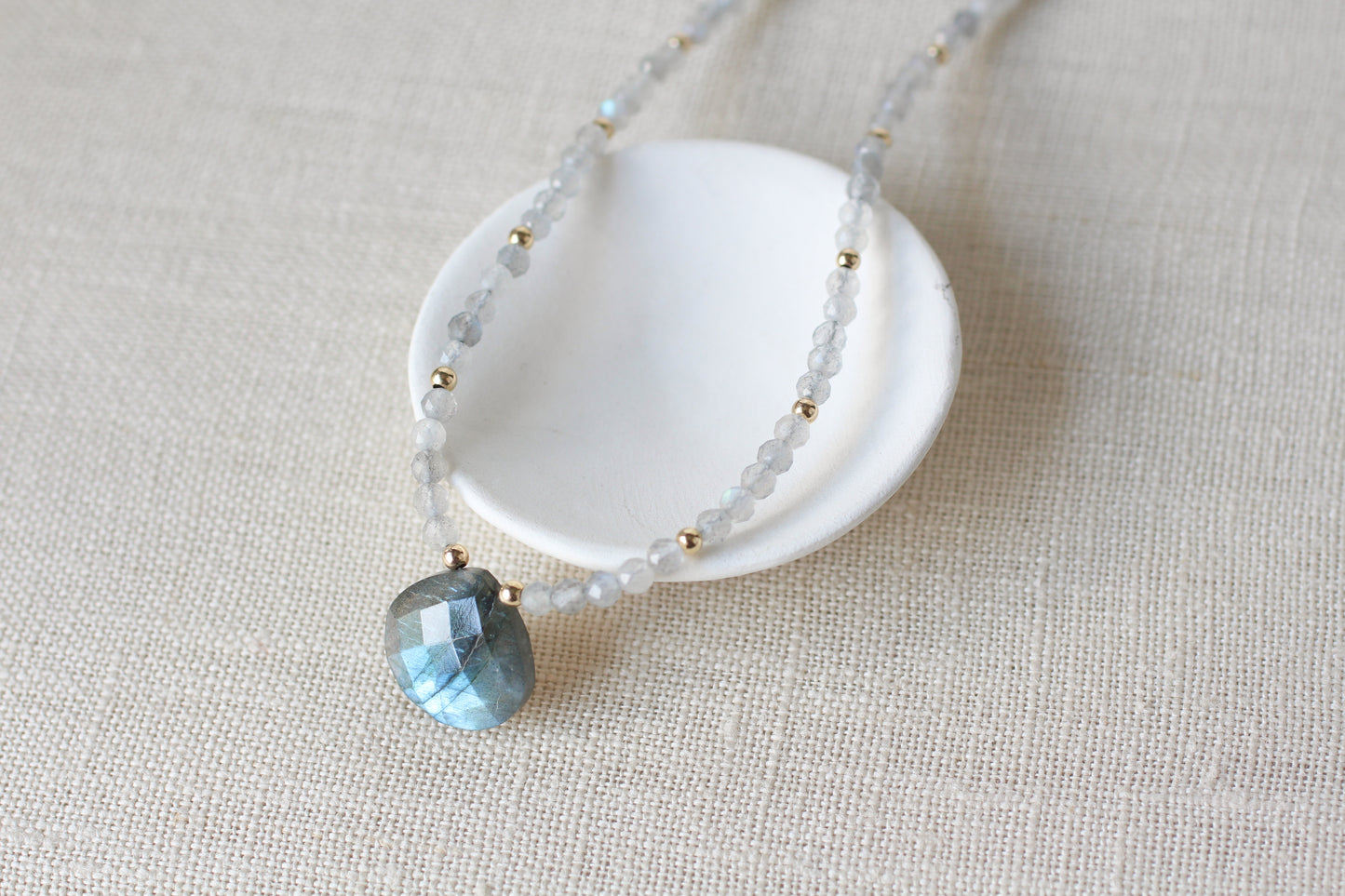 A delicate beaded necklace with pale blue-gray translucent beads, small gold spacer beads, and a faceted light blue teardrop pendant, displayed on a small white dish over a beige linen background.