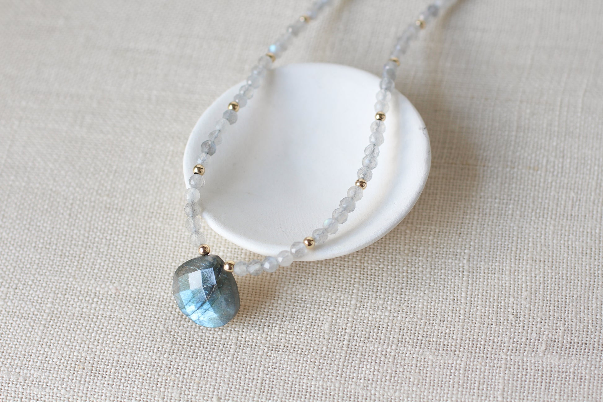 A delicate beaded necklace with pale blue-gray translucent beads, small gold spacer beads, and a faceted light blue teardrop pendant, displayed on a small white dish over a beige linen background.