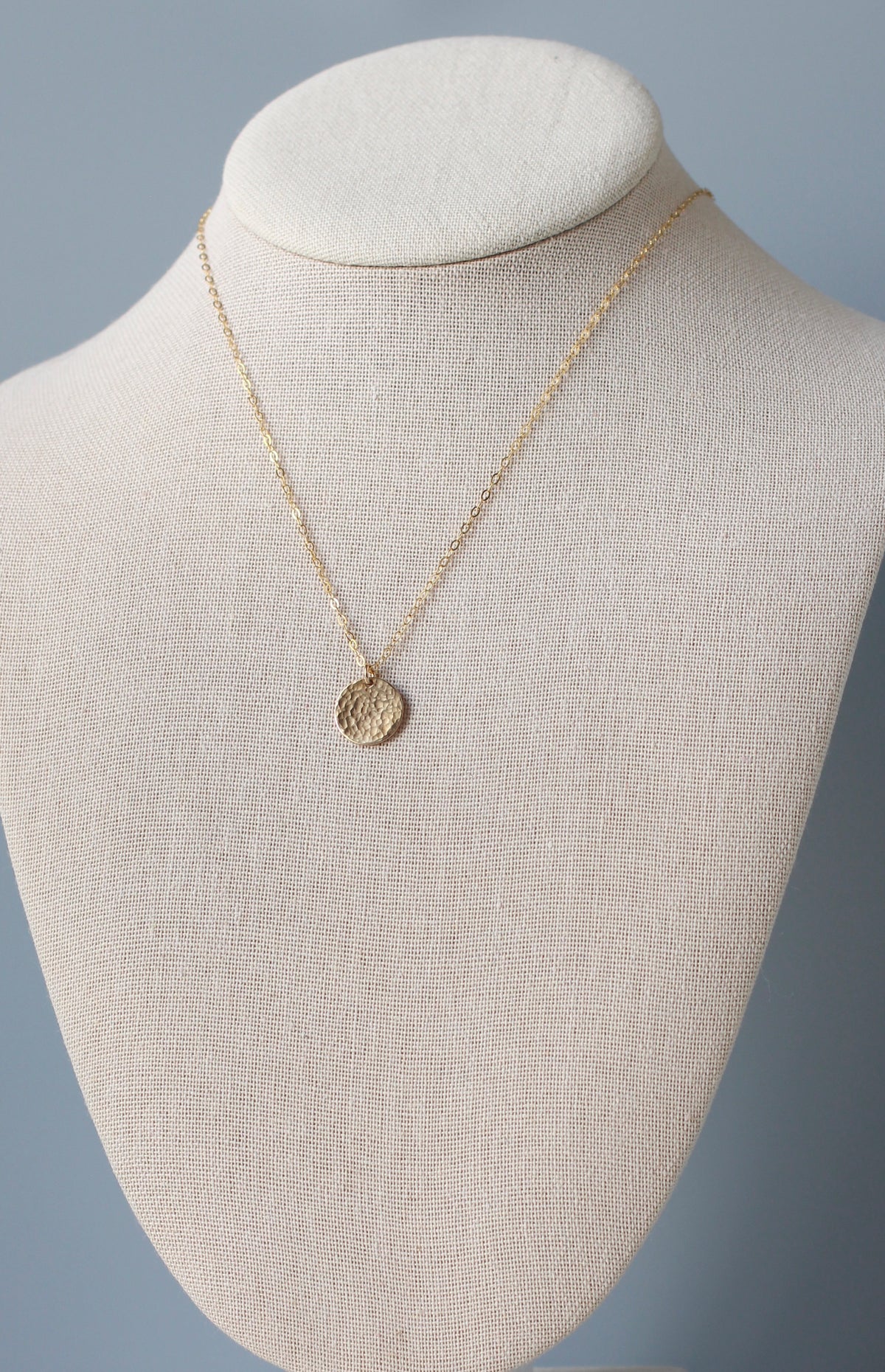 A delicate gold chain necklace with a small textured circular coin pendant, displayed on a beige fabric jewelry bust against a soft blue-gray background.