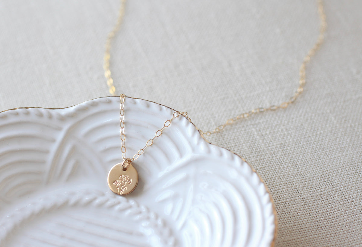 A delicate gold chain necklace with a small round gold disc pendant featuring an engraved carnation flower design, resting on a white ceramic dish over a textured beige linen background.