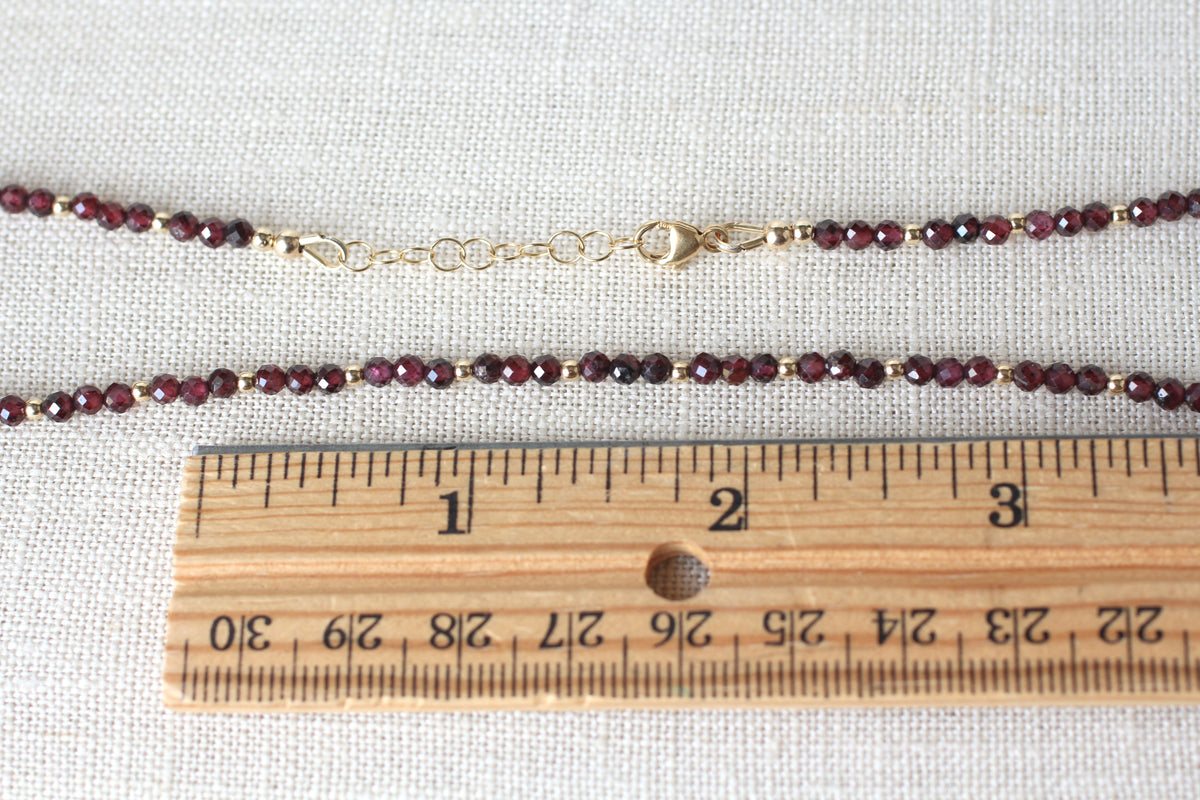 A beaded chain necklace with faceted dark red beads separated by small gold spacer beads and a gold clasp, shown on a light fabric background with a wooden ruler for scale.