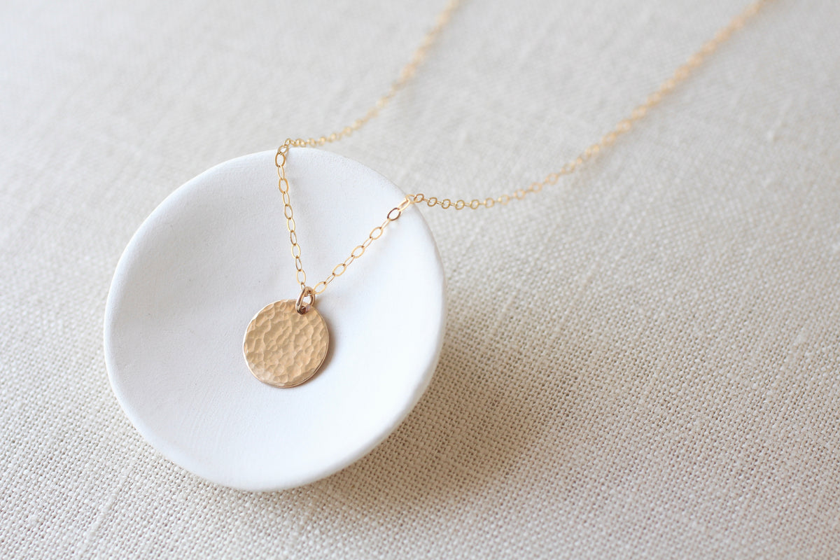 A delicate gold chain necklace with a small, textured circular gold pendant, displayed on a small white ceramic dish over a beige linen fabric background.