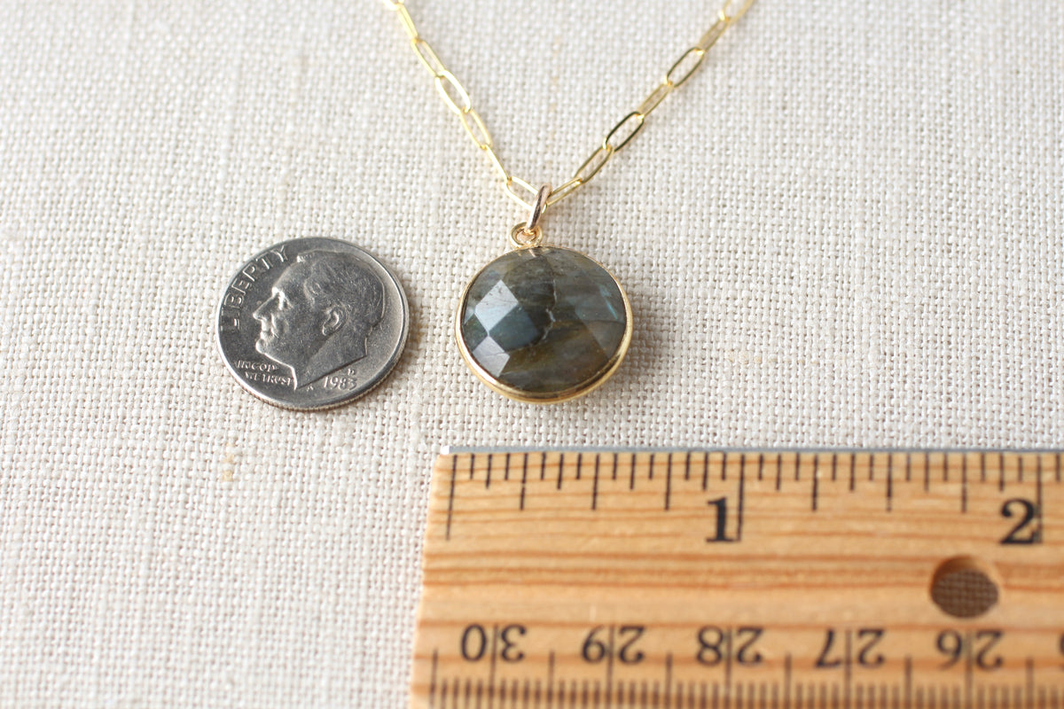 A gold chain necklace with a round faceted gray stone pendant, shown next to a U.S.